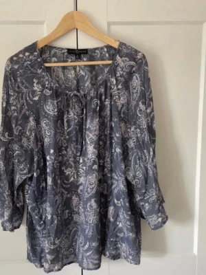 Paisleymönstrad blus  - Snygg blå blus fmed paisleymönster i vitt.. Blusen har rund halsringning med knytband och är tillverkad i ett tunt, luftigt material som ger en avslappnad look. Perfekt till jeans eller kjol. Storlek USA 1xl