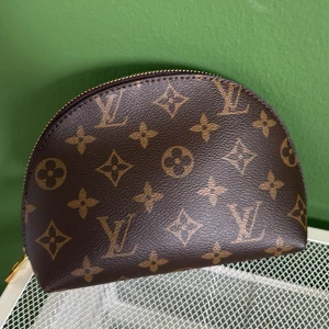 Brun sminkväska necessär monogram mönster - Säljer en klassisk halvmåneformad necessär i brunt läder med det ikoniska monogrammönstret i guld. Väskan har dragkedja upptill och är perfekt för smink eller småprylar. En riktig lyxdetalj till din väsksamling.