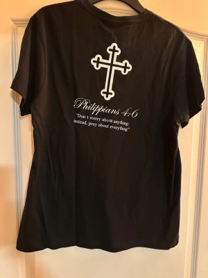Svart t-shirt med kors och citat - Svart t-shirt med vitt kors på bröstet och större kors på ryggen tillsammans med texten 'Philippians 4:6' och ett citat. Klassisk rund hals och korta ärmar. Tillverkad i mjuk bomull för en bekväm känsla.