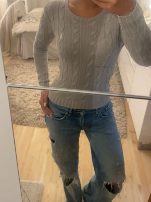 Ljusgrå kabelstickad tröja - Säljer en ljusgrå kabelstickad tröja från ralph lauren med rund halsringning och långa ärmar. Tröjan är figurnära och har klassiskt flätmönster framtill och på ärmarna. Nytt skick då jag använt den 2 gånger 