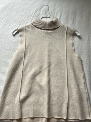 Beige ärmlös stickad poloväst - Säljer en stilren beige stickad väst med hög polokrage. Västen är ärmlös och har en rak, avslappnad passform. Perfekt att bära över en skjorta eller långärmad tröja för en trendig lager-på-lager-look.