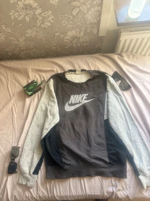 Nike sweatshirt i grå och svart - Snygg sweatshirt från Nike i grått och svart med stort vitt Nike-logotyptryck på bröstet. Tröjan har långa ärmar och rund halsringning. Perfekt för en sportig och avslappnad stil. Materialet känns mjukt och bekvämt.