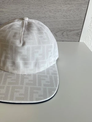 Vit keps med logga - Säljer en stilren vit keps med diskret monogram över hela kepsen. Klassisk böjd skärm och justerbar baksida. Tillverkad i ett tåligt tyg med svart insida och detaljer. Perfekt för dig som gillar exklusiva accessoarer.