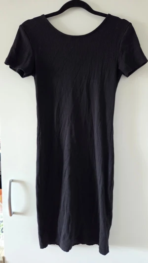 Svart ribbad kortklänning - En svart ribbad kortklänning med rund halsringning fram och djup urringning i ryggen. Klänningen har korta ärmar och är figurnära i modellen. Perfekt för dig som gillar enkel men snygg stil.