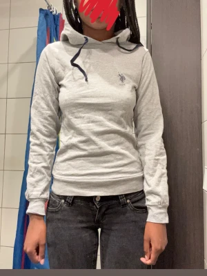 Grå hoodie med tight passform från U.S. Polo Assn. - Säljer en ljusgrå hoodie med tight passform från U.S. Polo Assn. med mörkblå dragsko i huvan.