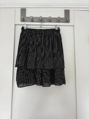 Svart rutig volangkjol - Säljer en svart kortkjol med vitt rutmönster och volanger. Kjolen har resår i midjan och är tillverkad i ett lätt, flödande material som ger en skön och luftig känsla. Perfekt för dig som gillar en trendig och avslappnad stil. Gissar på stlk S
