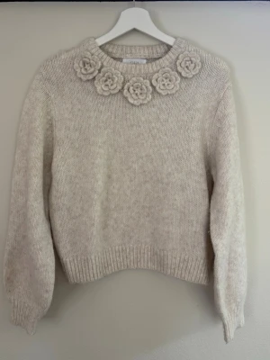 Beige stickad tröja med blommor Zara - Supersöt beige stickad tröja från Zara med stora virkade blommor runt halsringningen. Tröjan har rund hals, långa ärmar och ribbade muddar. Perfekt för dig som gillar unika detaljer och mjuka material.