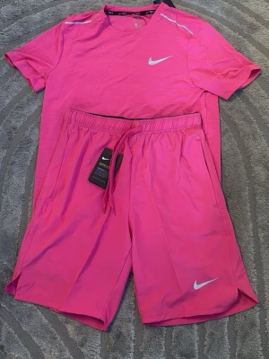Rosa träningsset från Nike - Snyggt rosa träningsset från Nike med t-shirt och shorts. T-shirten har rund hals, korta ärmar och diskreta ränder i tyget samt vit Nike-logga på bröstet. Shortsen har resårmidja med dragsko och vit logga på benet. Perfekt för gymmet eller löprundan.