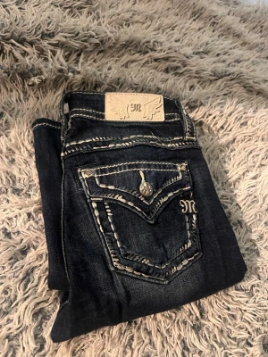 Miss Me jeans  - Säljer ett par äkta Miss Me Mid-Rise Boot jeans i storlek W26 (L32).  Style: M5014B406V  Detta är en äldre Miss Me-modell som inte längre tillverkas, vilket gör den svår att hitta idag. Perfekt för dig som gillar den klassiska Y2K-stilen.  Byxorna är i nästan nyskick och har endast använts 2–3 gånger innan de blev liggande i garderoben. Inga hål, fläckar eller slitage.   Mörk tvätt med de ikoniska vita sömmarna och de klassiska bakfickorna. 