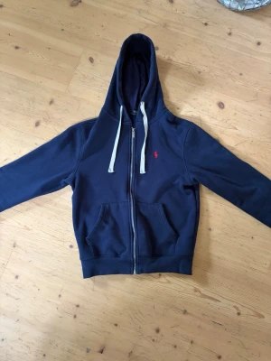 Marinblå hoodie från Polo Ralph Lauren - Snygg marinblå hoodie från Polo Ralph Lauren med dragkedja, vit dragsko och klassisk röd logga på bröstet. Tillverkad i mjuk bomullsblandning och har två fickor framtill. Perfekt för en avslappnad och stilren look.