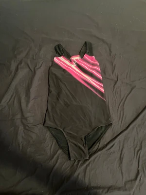Svart baddräkt med rosa mönster - Sportig svart baddräkt från SOC med breda axelband och ett diagonalt rosa och vitt mönster framtill. Baddräkten har en klassisk rygg med stor öppning och är tillverkad i stretchigt material som passar perfekt för simning och träning.
