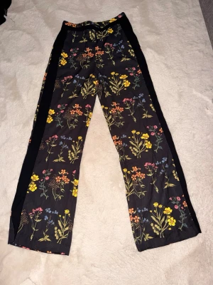 Svarta byxa med blommigt mönster - Snygga svarta byxa med färgglatt blommönster i gult, rosa och blått. Byxan har raka ben och svarta sidopaneler som ger en sportig touch. Perfekta för dig som vill sticka ut med en unik och trendig stil.