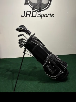 Golfset med bag från Spalding - Komplett golfset från Spalding med svart golfbag och flera klubbor, inklusive järn, driver och putter. Bagen har vita detaljer och flera fack för förvaring. En av dragkedjorna på bagen är trasig. Perfekt för dig som vill komma igång med golf eller uppgradera din utrustning.