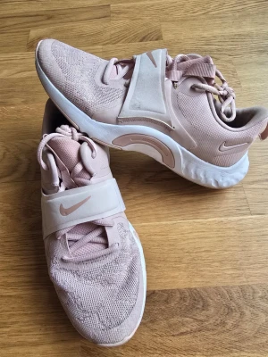Ljusrosa Nike sneakers med rem - Snygga ljusrosa sneakers från Nike med stickad ovandel och bred rem över foten. Skorna har vita sulor och klassisk Nike-logga på sidan och remmen. Perfekta för dig som gillar sportig och trendig stil.