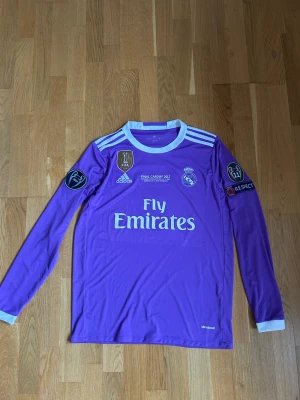 Lila Real Madrid Ronaldo tröja - Säljer en lila långärmad Real Madrid matchtröja från Adidas med Ronaldo 7 på ryggen. Tröjan har vita detaljer, Champions League och FIFA-märken på ärmarna samt Fly Emirates-tryck på bröstet. Perfekt för dig som älskar fotboll och vill sticka ut på planen.