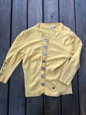 Gul kofta från Karen Millen - Snygg gul kofta från Karen Millen med stora dekorativa knappar framtill och ribbade muddar. Koftan har rund halsringning, trekvartsärmar och fina knappdetaljer på ena ärmen. Perfekt för dig som vill ha en färgklick i garderoben.