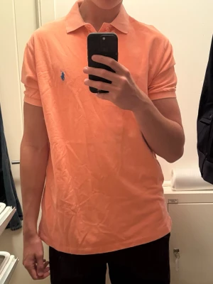 Persikofärgad piké från Ralph Lauren - Säljer en persikofärgad pikétröja från Ralph Lauren med klassisk krage och korta ärmar. Tröjan har storlek L men känns som XL. Tröjan har den ikoniska blå logotypen broderad på bröstet och är tillverkad i mjuk bomull. Perfekt för en clean och stilren look.
