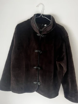 Mörkbrun pälsjacka med knäppning - Mysig mörkbrun pälsjacka i fuskpäls med hög krage och dekorativa knappar framtill. Jackan har en rak passform och är fodrad för extra värme. 