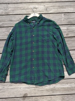 Grönblå rutig Flannel skjorta från Chaps - Klassisk grön och blå rutig skjorta från Chaps med knappar framtill och bröstficka. Skjortan har lång ärm och en avslappnad passform, perfekt för en chill och stilren look. Tillverkad i mjuk bomull som känns skön mot huden.