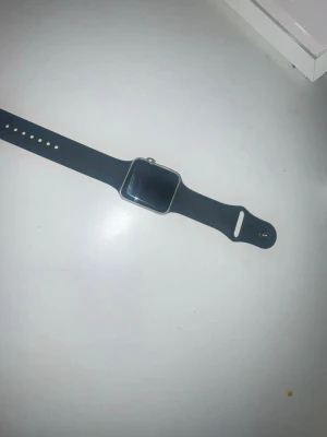 Apple Watch med svart sportarmband - Snygg Apple Watch med svart sportarmband i silikon och fyrkantig aluminiumboett. Klockan har en stilren design med digital skärm och klassiskt spänne. Perfekt för dig som vill ha koll på notiser, träning och hälsa direkt på handleden.