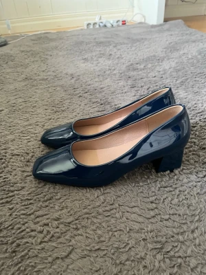 Mörkblå lackpumps med klack - Snygga mörkblå pumps i lackmaterial med fyrkantig tå och bred klack. Skorna har en stilren och modern look med slät yta och beige innersula. Perfekta för dig som vill ha en klassisk men trendig sko.