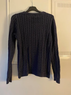 Mörkblå kabelstickad tröja - Mörkblå kabelstickad tröja med rund halsringning och liten logga på bröstet. Tröjan har långa ärmar och ribbade muddar vid ärmslut och nederkant. Perfekt för lager på lager och en klassisk look.