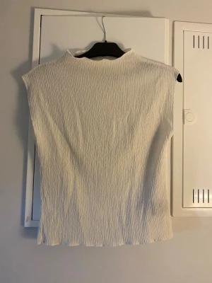 Vit ribbad topp med hög hals - Säljer en stilren vit topp med hög hals och ribbad struktur. Toppen är ärmlös och har en lätt croppad passform som ger en modern look. Perfekt att matcha med jeans eller kjol för en trendig outfit.