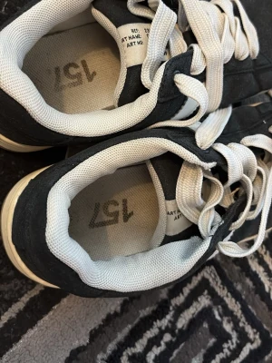 Svarta och vita sneakers med snörning - Snygga sneakers i svart och vitt med klassisk snörning och vit sula. Ovandelen är i textil och syntet, med vadderad kant och meshfoder för extra komfort. Storlek US 5. Passar perfekt till en avslappnad streetstyle.