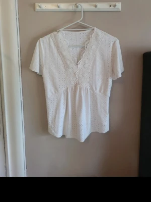 Vit blus med spets och broderi - Söt vit blus med v-ringning, spetsdetaljer och broderade hålmönster. Blusen har korta ärmar och lätt rynkad midja för en feminin siluett. Tillverkad i ett tunt, luftigt bomullstyg som passar perfekt till sommaren.