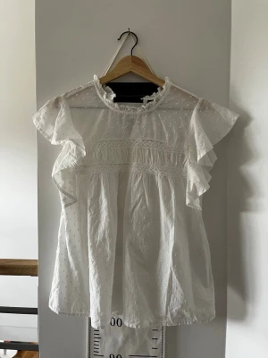 Vit volangblus med spetsdetaljer - Söt vit blus med volangärmar och transparenta partier upptill. Blusen har fina spetsdetaljer och diskret prickmönster i tyget. Luftig och flowy passform, perfekt för en trendig och romantisk look. Rundad halsringning med liten volangkant. Använd en gång