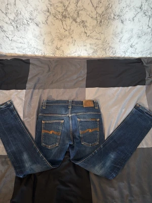 Nudie Jeans - Säljer ett par blå slim fit jeans från Nudie Jeans med klassiska kontrastsömmar och slitningar framtill. Jeansen har fem fickor och Nudie Jeans signaturbroderi på bakfickorna. Tillverkade i denim med snygg tvätt och läderpatch i midjan.