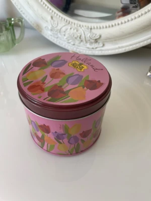 Rund plåtburk med tulpanmotiv - Säljer en rund plåtburk i rosa med färgglada tulpaner och texten 'Holland' på locket. Perfekt för småförvaring eller som dekoration. Burken har ett tättslutande lock och är tillverkad i metall.