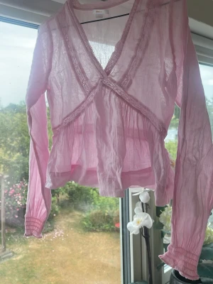 Rosa transparent blus från Gina Tricot - Söt rosa blus från Gina Tricot i transparent, tunn och luftig kvalitet. Blusen har omlott-skärning med spetsdetaljer längs kanterna, volang nedtill och långa ärmar med resår vid ärmsluten. Perfekt för en trendig och feminin look.