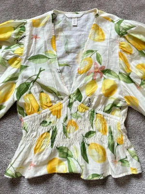 Blommig citrontopp med puffärm - Superfin topp från H&M med citrongult och grönt blommönster på vit botten. Djup v-ringning, puffiga trekvartsärmar och smockad midja som ger en snygg siluett. Perfekt för sommarens alla tillfällen! Toppen är aldrig använd tyvärr så o helt nytt skick💛