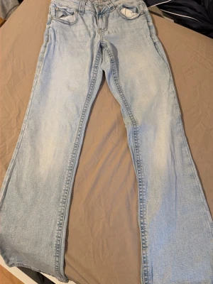 Ljusblå utsvängda jeans - Säljer ett par ljusblå jeans med utsvängda ben och klassisk femficksdesign. Jeansen har en avslappnad passform och är tillverkade i denim. Perfekta för en trendig och avslappnad stil.