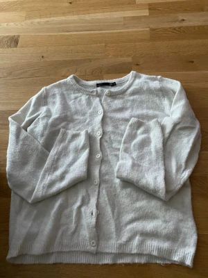 Vit kofta från 157, rund hals - Mysig vit kofta från 157 med rund halsringning och knäppning framtill. Koftan har långa ärmar och ribbade muddar vid ärmslut och nederkant. Perfekt att slänga över en t-shirt eller linne för en enkel och clean look.