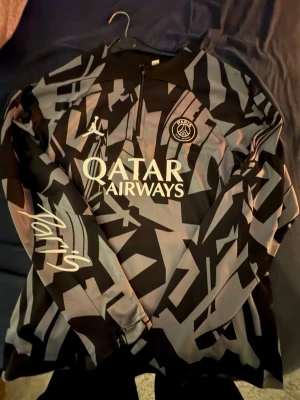 PSG träningströja svart/grå mönstrad - Säljer en svart och grå mönstrad träningströja från PSG i samarbete med Jordan. Tröjan har halv dragkedja, lång ärm och tryck med Qatar Airways på bröstet samt PSG-logga. Materialet är troligtvis polyester, perfekt för träning eller uppvärmning.