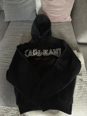 Svart Karl Kani hoodie med tryck - Säljer en svart hoodie från Karl Kani med stor logga och texten 'EST. 1989' på bröstet. Klassisk modell med huva och dragsko, samt ribbade muddar vid ärmar och midja. Perfekt för en avslappnad passform. Storleken på tröjan är S men passar lika bra på folk med storlek M.
