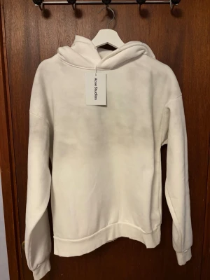 Vit hoodie från Acne Studios - Vit hoodie från Acne Studios med tryck på ryggen där det står 'Acne Studios STOCKHOLM 1996'. Klassisk passform med huva och ribbade muddar. Perfekt för en clean och stilren look.