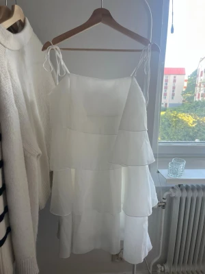 Vit volangklänning med knytband - Säljer en söt vit kortklänning med tunna axelband som knyts på axlarna. Klänningen har flera lager volanger och är tillverkad i ett lätt, luftigt material som passar perfekt till varma dagar. Enkel och stilren design med romantisk känsla. Oanvänd. Nypris: 699kr 