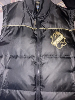 Svart dunväst från AIK - Snygg svart dunväst från AIK med gulddetaljer och klubbens emblem broderat på bröstet. Västen har dragkedja framtill och broderad text '1891' på ryggen. Perfekt för dig som vill visa ditt AIK-stöd med stil.