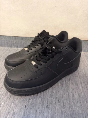 Svarta Nike Air Force 1 sneakers - Klassiska helsvarta Nike Air Force 1 sneakers i lågt utförande. Skorna har ovandel i slätt läder, perforerad tåbox och svart sula. Swoosh-logga på sidan och svarta skosnören med metall-detalj framtill. Perfekta för en clean och stilren look.