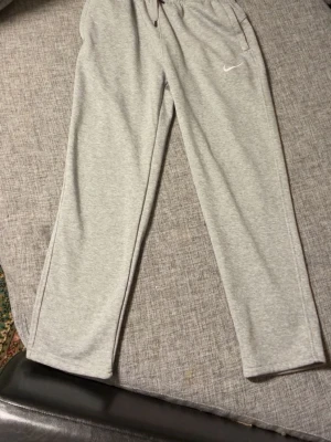 Grå mjukisbyxor från Nike - Snygga ljusgrå mjukisbyxor från Nike med klassisk vit swoosh-logga på benet. Byxorna har raka ben, resår i midjan med dragsko och sidofickor. Perfekta för chill eller träning, tillverkade i mjukt och bekvämt material.
