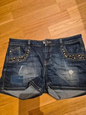 Mörkblå jeansshorts med nitar - Säljer ett par mörkblå jeansshorts med guldfärgade nitar längs fickorna fram. Shortsen har slitna detaljer, patch och uppvikta benslut för en cool och avslappnad look. Perfekta för sommaren och ger en edgy vibe till din outfit.