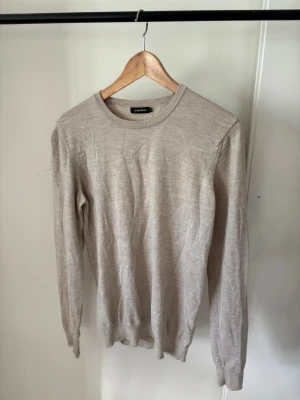 J.Lindeberg tröja - Snygg beige stickad tröja med rund halsringning och långa ärmar. Tröjan är tunn och har ribbade muddar vid ärmslut och nederkant. Perfekt för lager på lager och enkel att matcha med olika outfits.