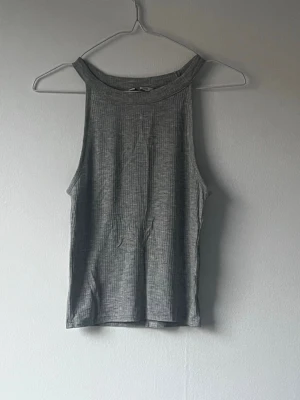 Grå ribbad ärmlös linnetopp - Snygg grå ribbad linnetopp med hög halsringning och ärmlös design. Perfekt till jeans eller kjol för en clean och enkel look. Materialet är mjukt och stretchigt för skön passform.
