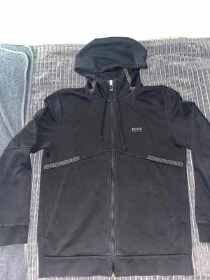 Äkta Hugo boss kofta - Snygg svart hoodie från BOSS med gulddetaljer på dragkedjan och logga på bröstet. Hoodien har hel dragkedja, justerbar huva med snören och diskreta mönsterdetaljer vid fickorna och huvan. Perfekt för en clean och stilren look.
