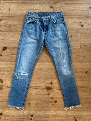 Levis 501 26” - Säljer ett par blå Levis 501  jeans med rak passform, slitningar på benen och råa, fransiga benslut. Jeansen har klassisk femficksdesign och knappgylf. Perfekta för en avslappnad och trendig look. Strl 26” 