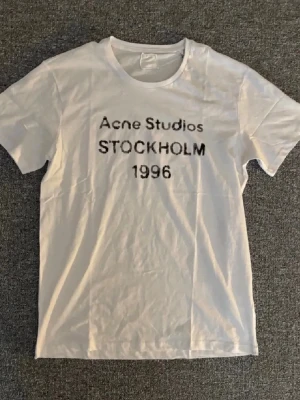 Vit Acne Studios t-shirt Stockholm - Vit t-shirt från Acne Studios med svart text 'Acne Studios STOCKHOLM 1996' på bröstet. Klassisk rund hals och korta ärmar. Tillverkad i mjuk bomull för en clean och stilren look.