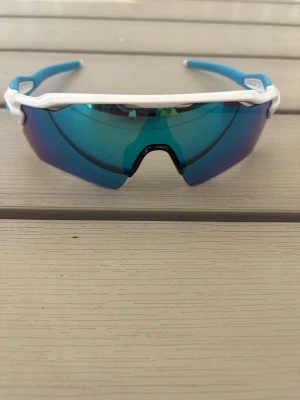 Oakley sportiga blå solglasögon - Säljer ett par sportiga solglasögon från Oakley med vit båge och blå spegelglas. De har en modern, svepande form och är perfekta för dig som gillar en aktiv livsstil. Snygg logga på sidan och bekväma skalmar i blått.